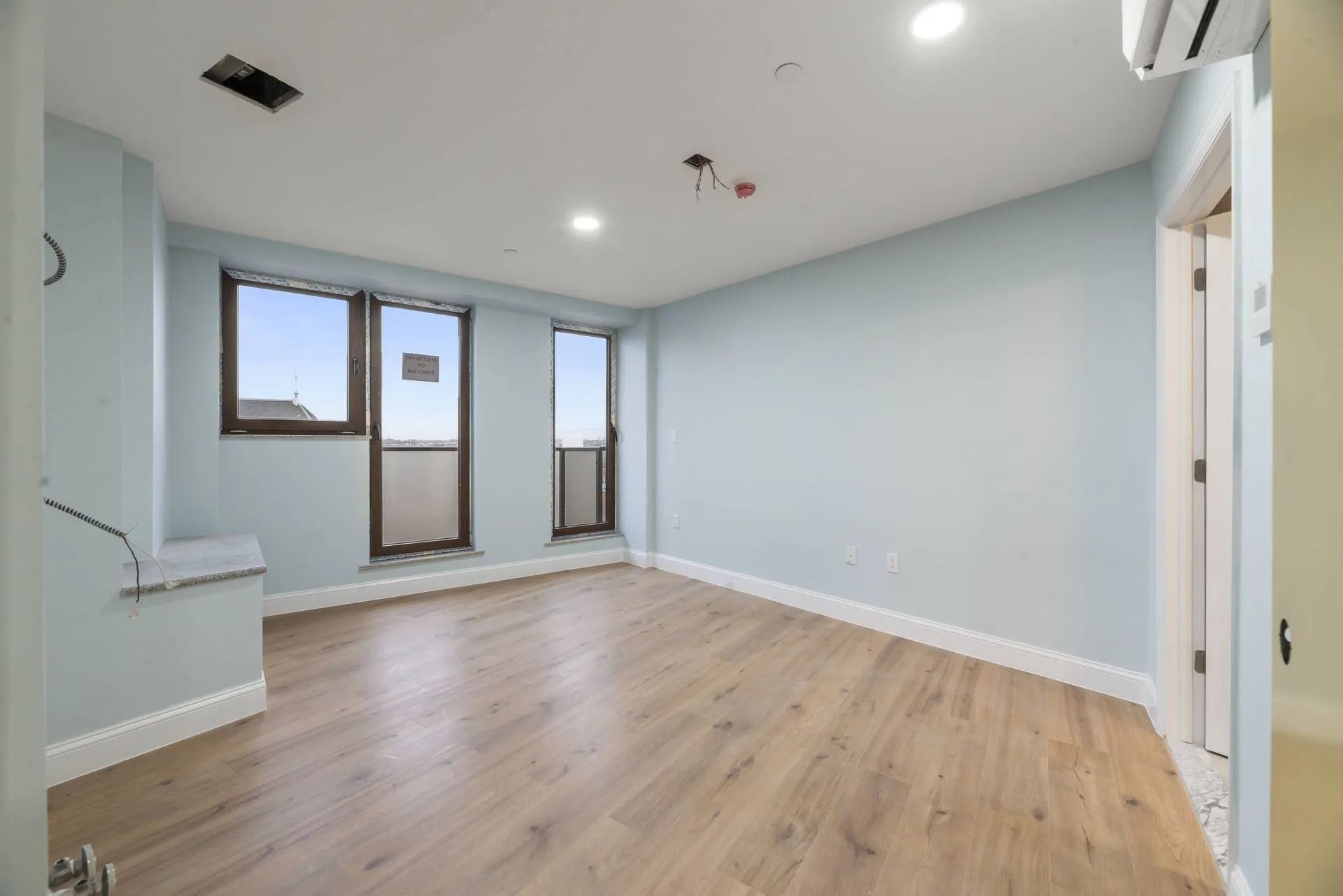Empty modern room with light wood floors, pale blue walls, recessed lighting, and large windows.