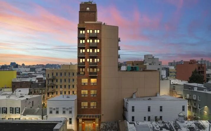 City skyline at sunset with a tall brick building lit by warm windows under a pink-blue sky