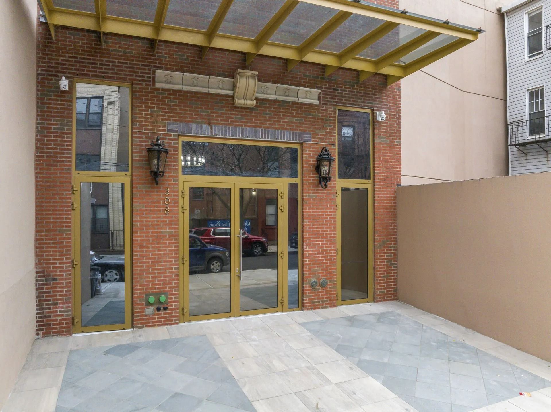 Brick building entrance with gold-framed glass doors, flanked by tall windows and a small paved forecourt.