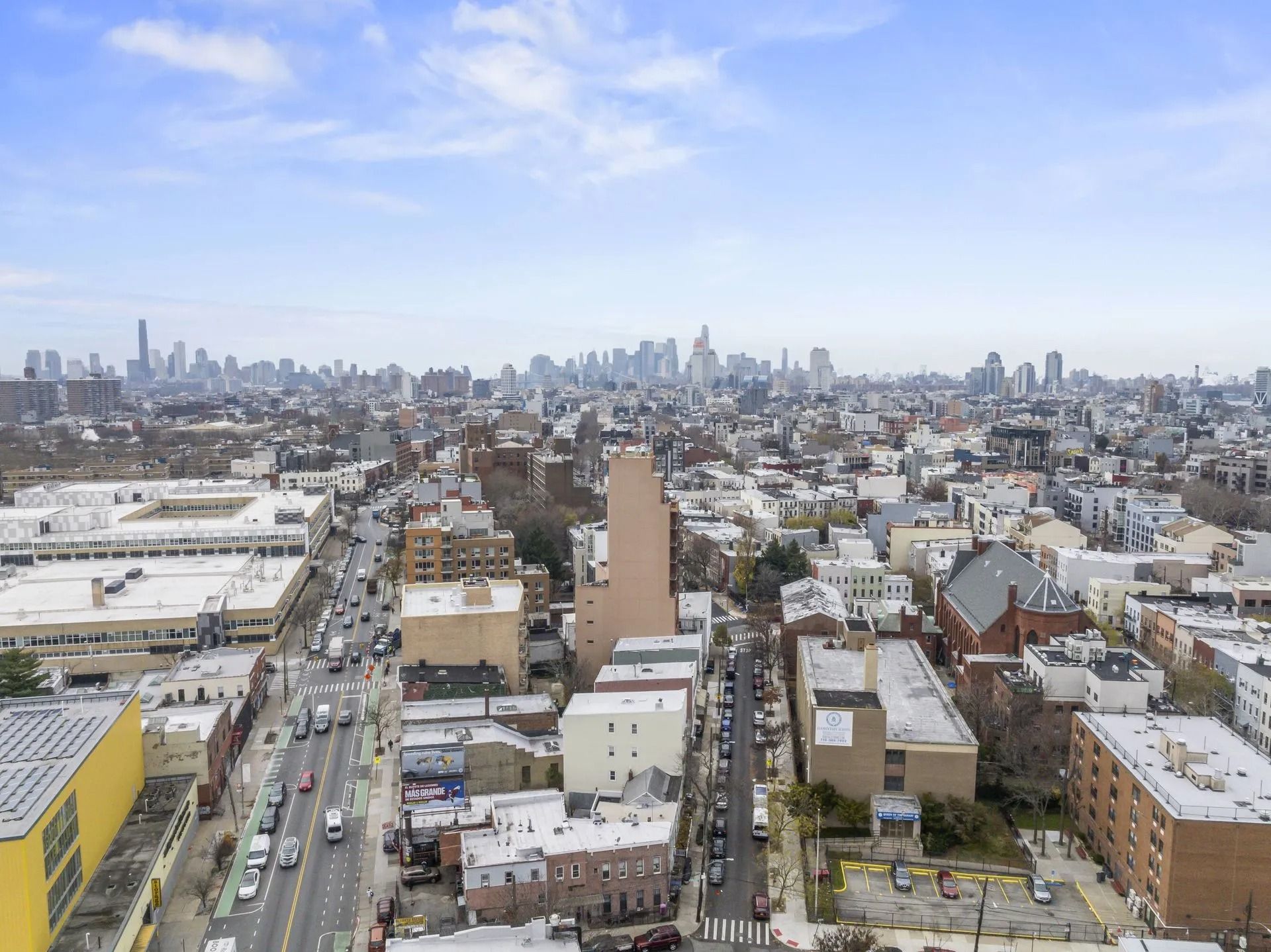 Aerial view of a dense city skyline under a partly cloudy blue sky, with streets and low-rise buildings in the foreground