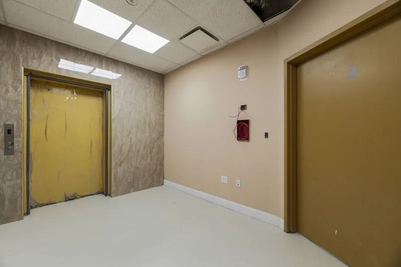 Empty hallway with beige walls, yellow double elevator doors, and a brown door on the right.