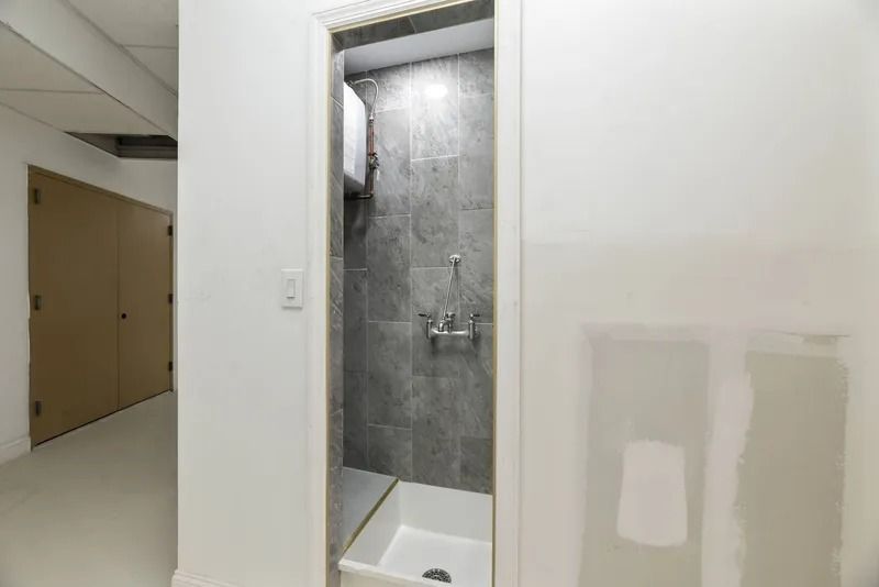 Small bathroom shower alcove with gray tile and white walls, next to a hallway and lockers