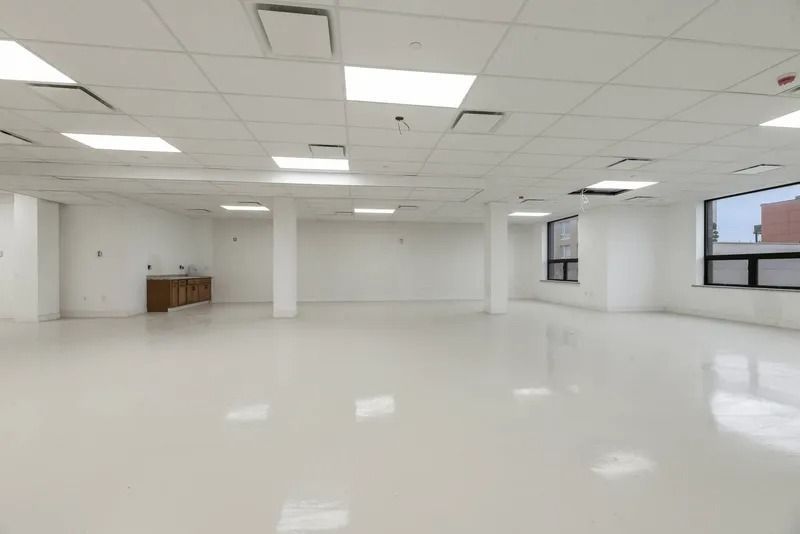 Empty white office space with glossy floor, ceiling lights, and windows along the right wall.