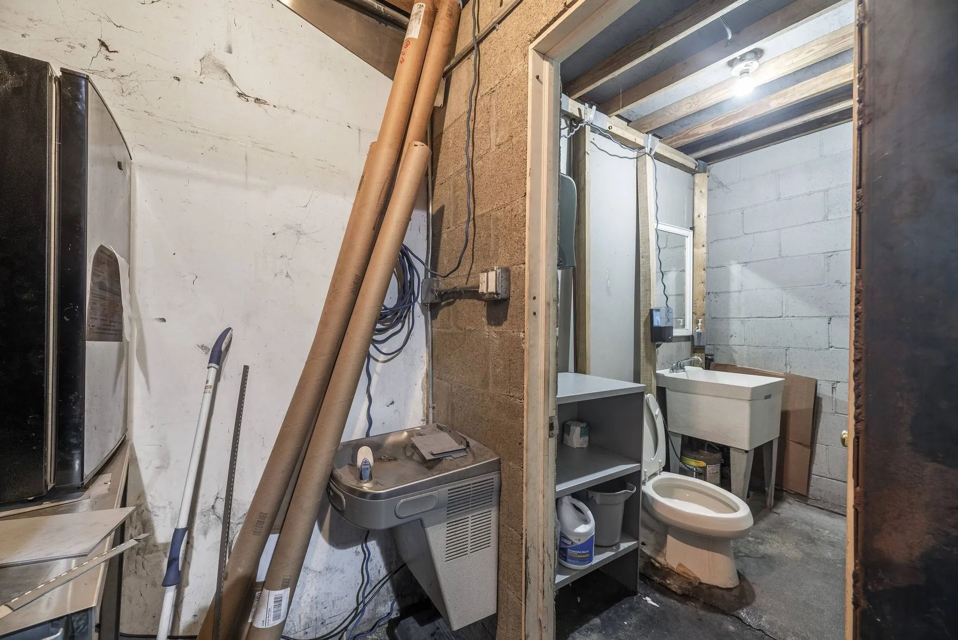 Cluttered unfinished basement bathroom with exposed pipes, sink, toilet, and storage shelves