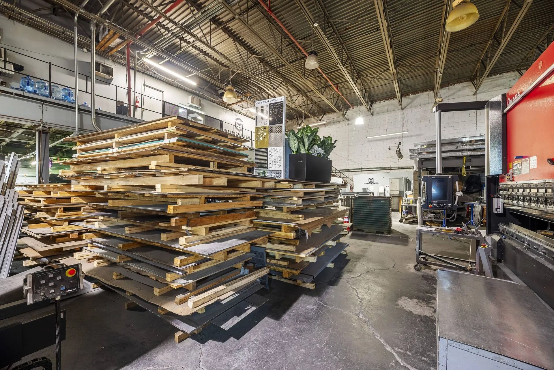 Stacked wooden boards in a bright industrial workshop with machinery and shelving.