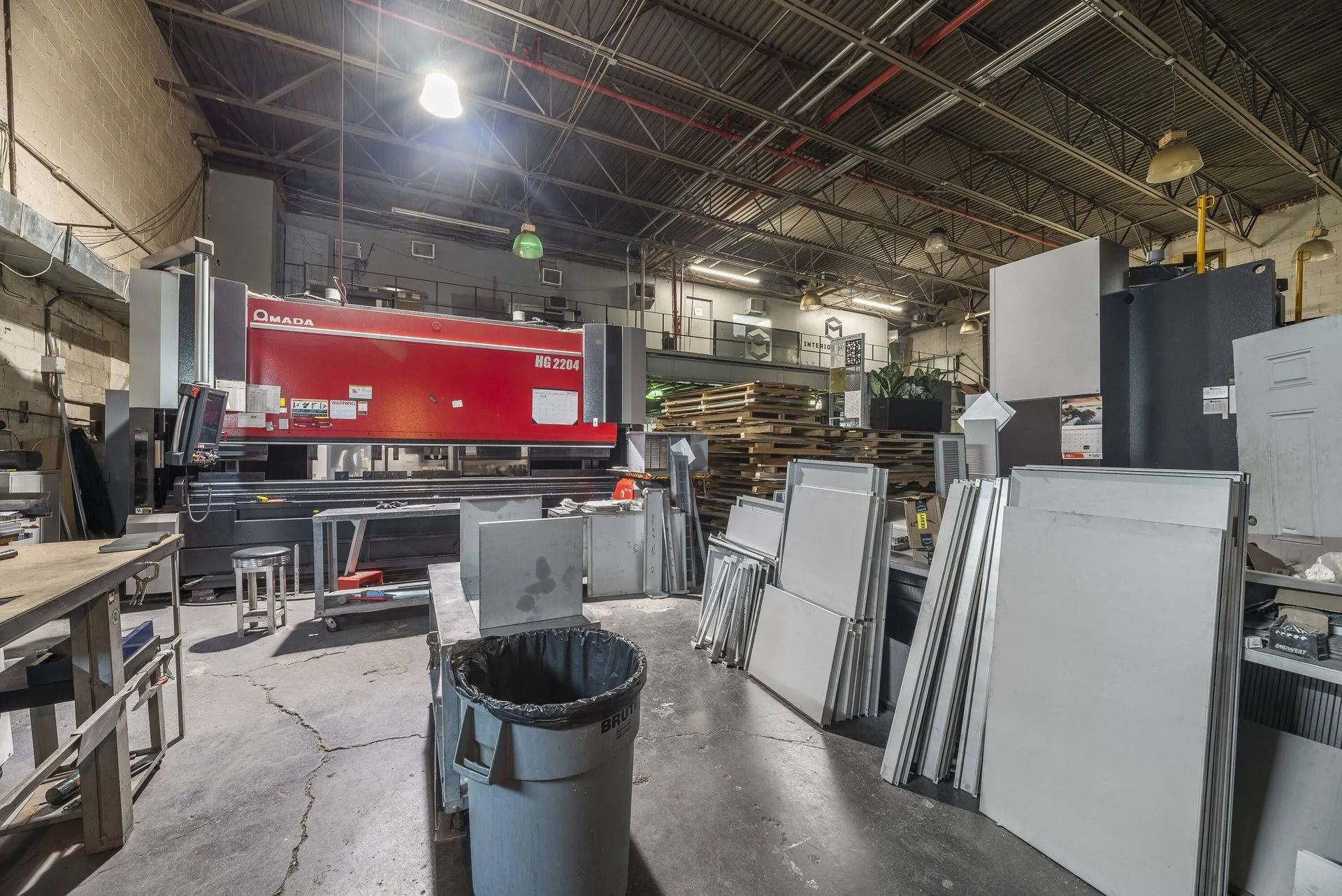 Industrial workshop with a large red machine, stacked panels, and tools under a high metal roof