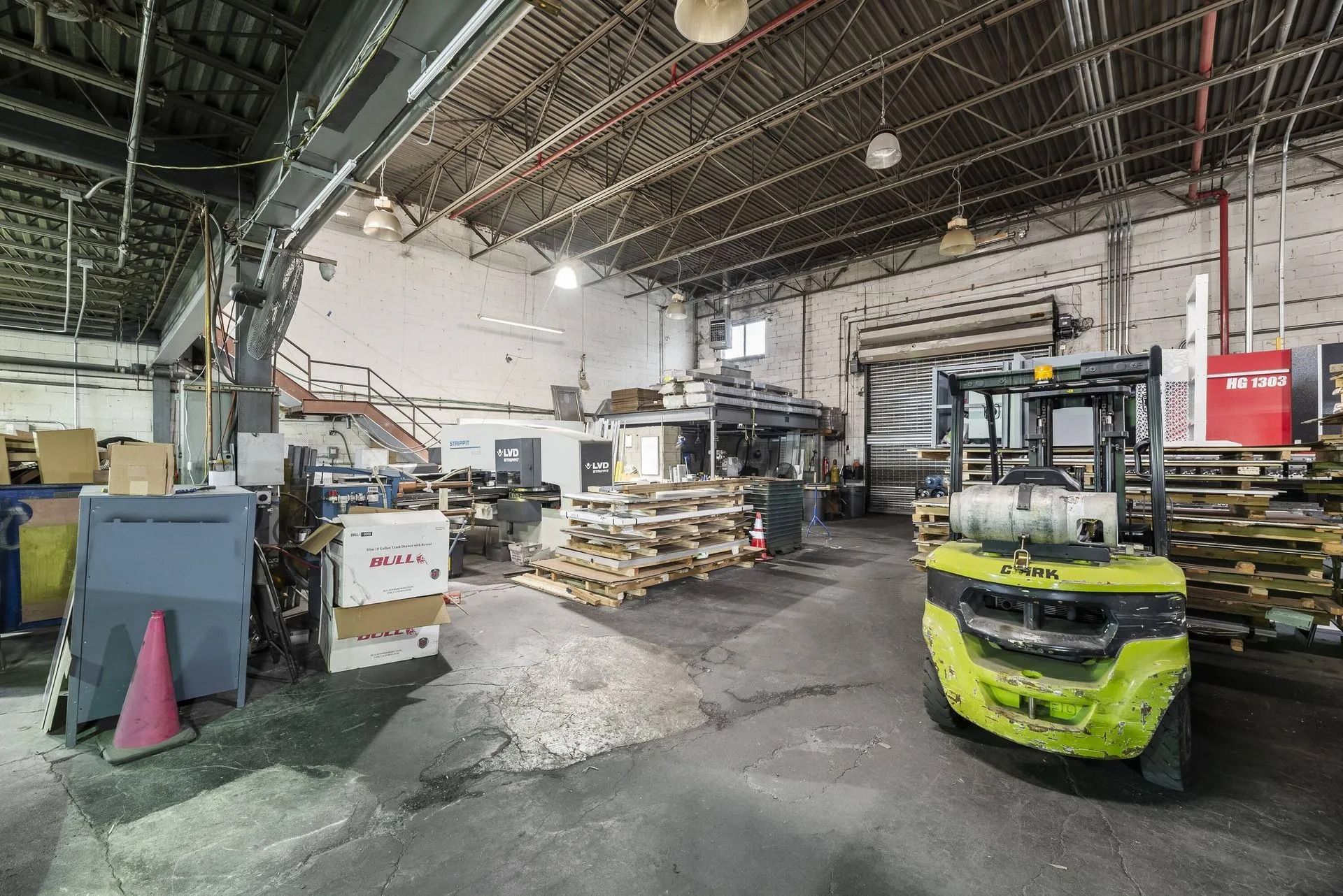 Indoor warehouse with a green forklift, stacked pallets, and industrial equipment under a high ceiling