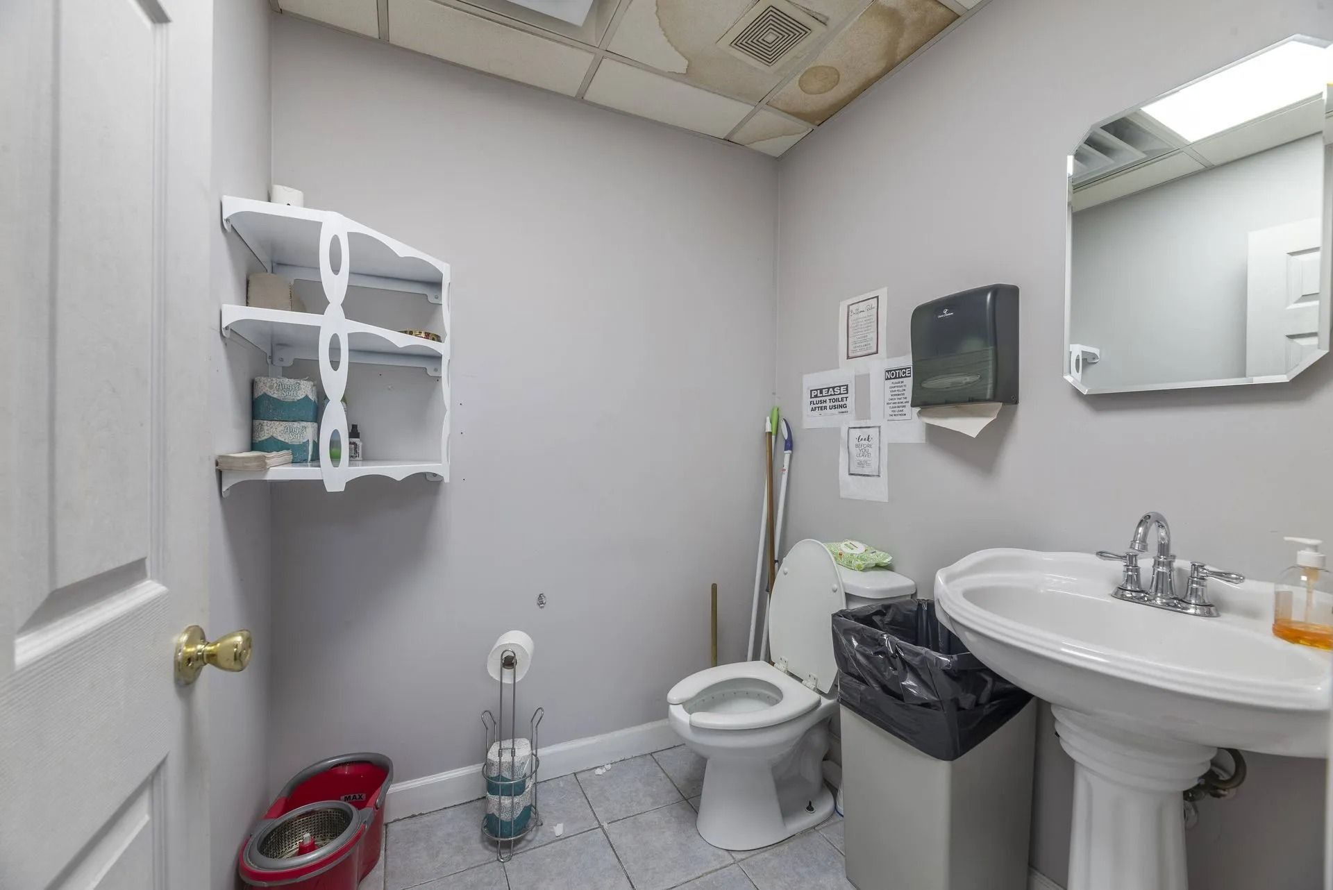 Small bathroom with sink, toilet, mirror, wall shelves, and cleaning supplies.