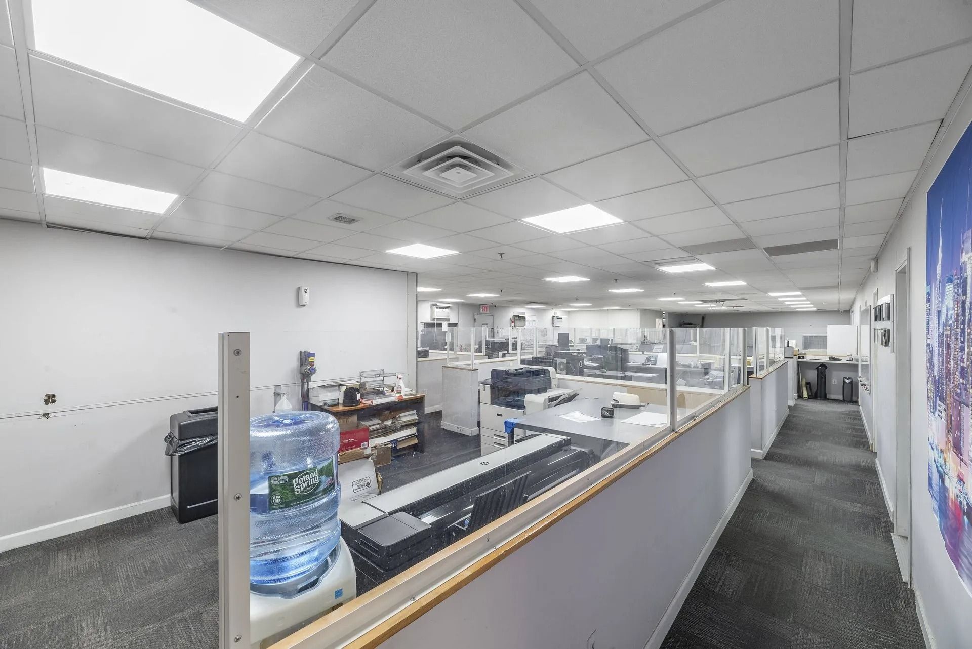 Bright office corridor with glass cubicles, desks, and a water cooler under fluorescent lights
