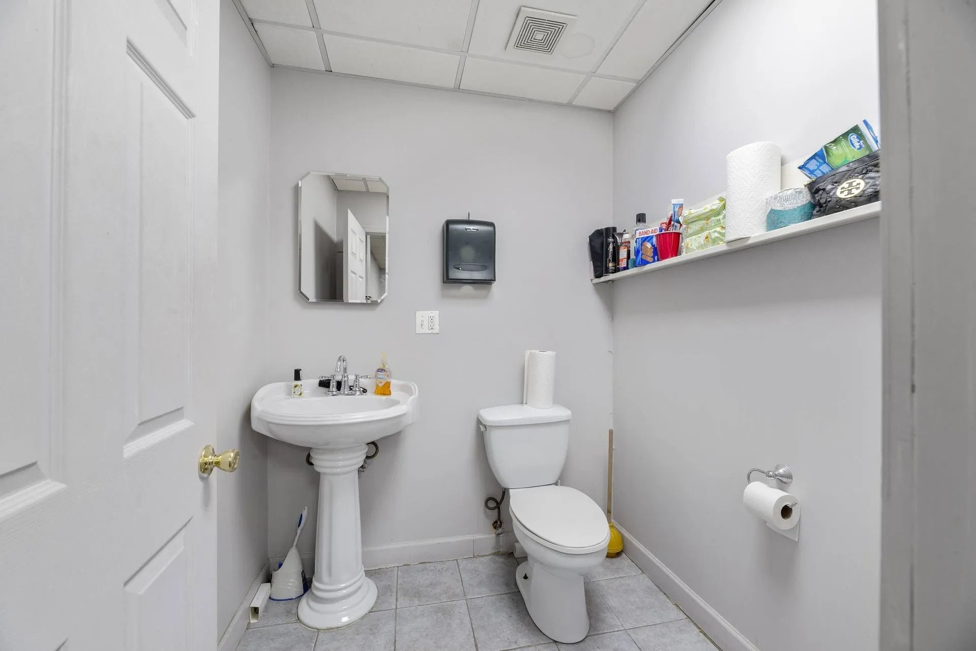 Small white bathroom with pedestal sink, toilet, mirror, and wall shelf.