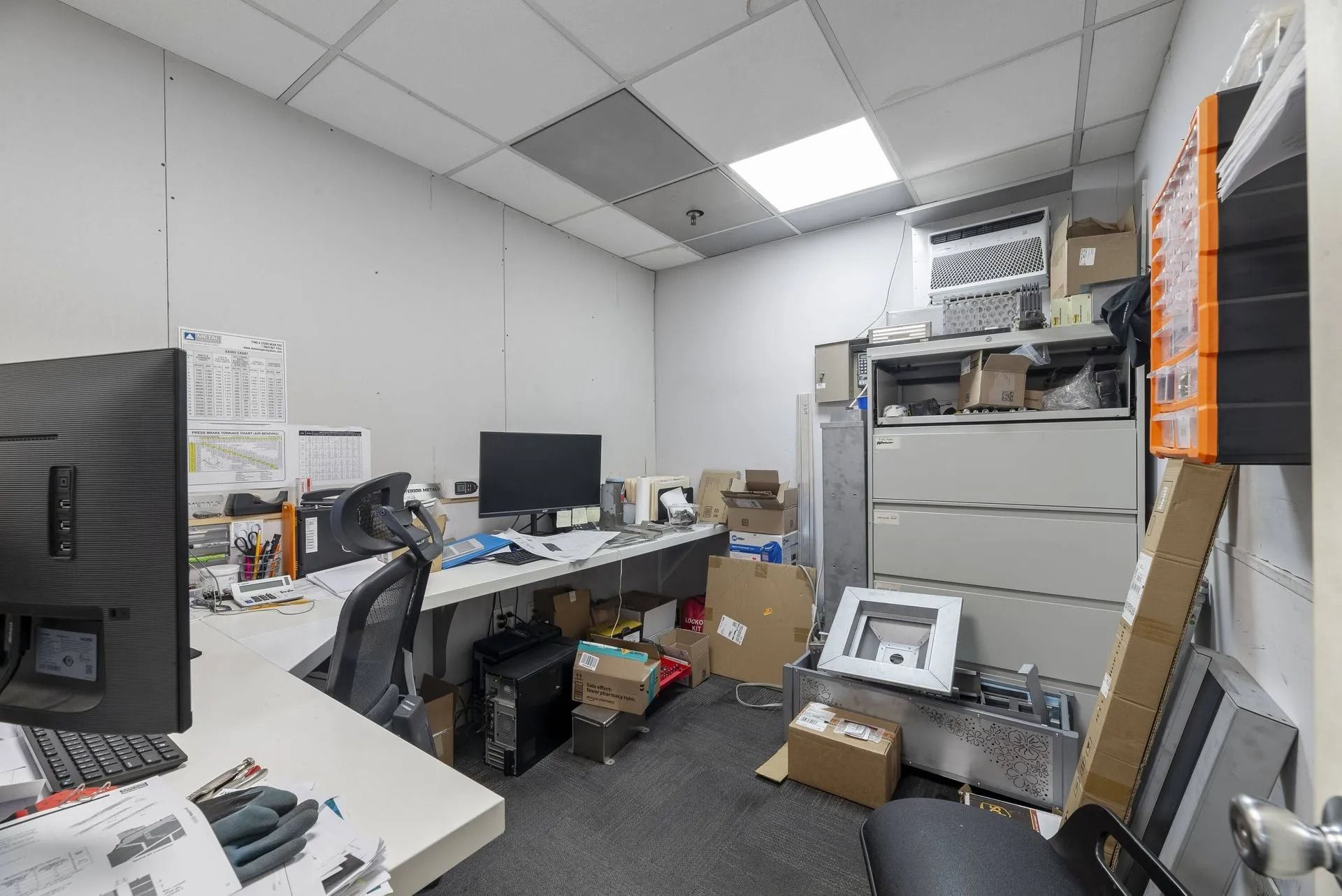 Cluttered office storage room with desks, monitors, cardboard boxes, and stacked filing cabinets.