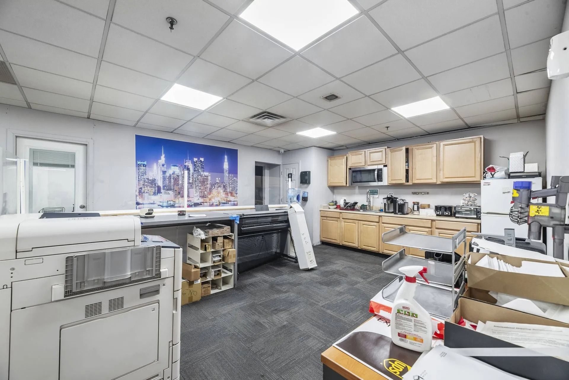 Office break room with desks, cabinets, and supplies under bright fluorescent lights