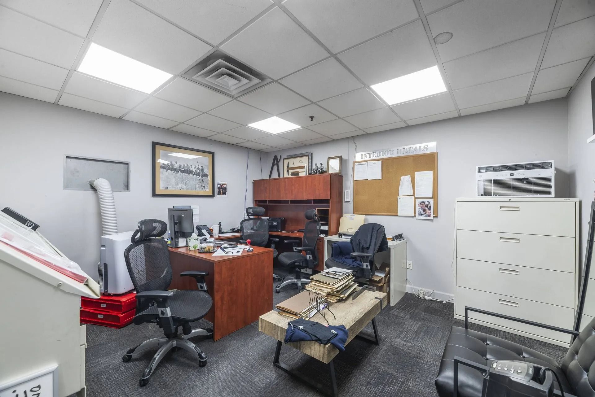 Office with desks, chairs, filing cabinets, and a corkboard under bright ceiling lights.