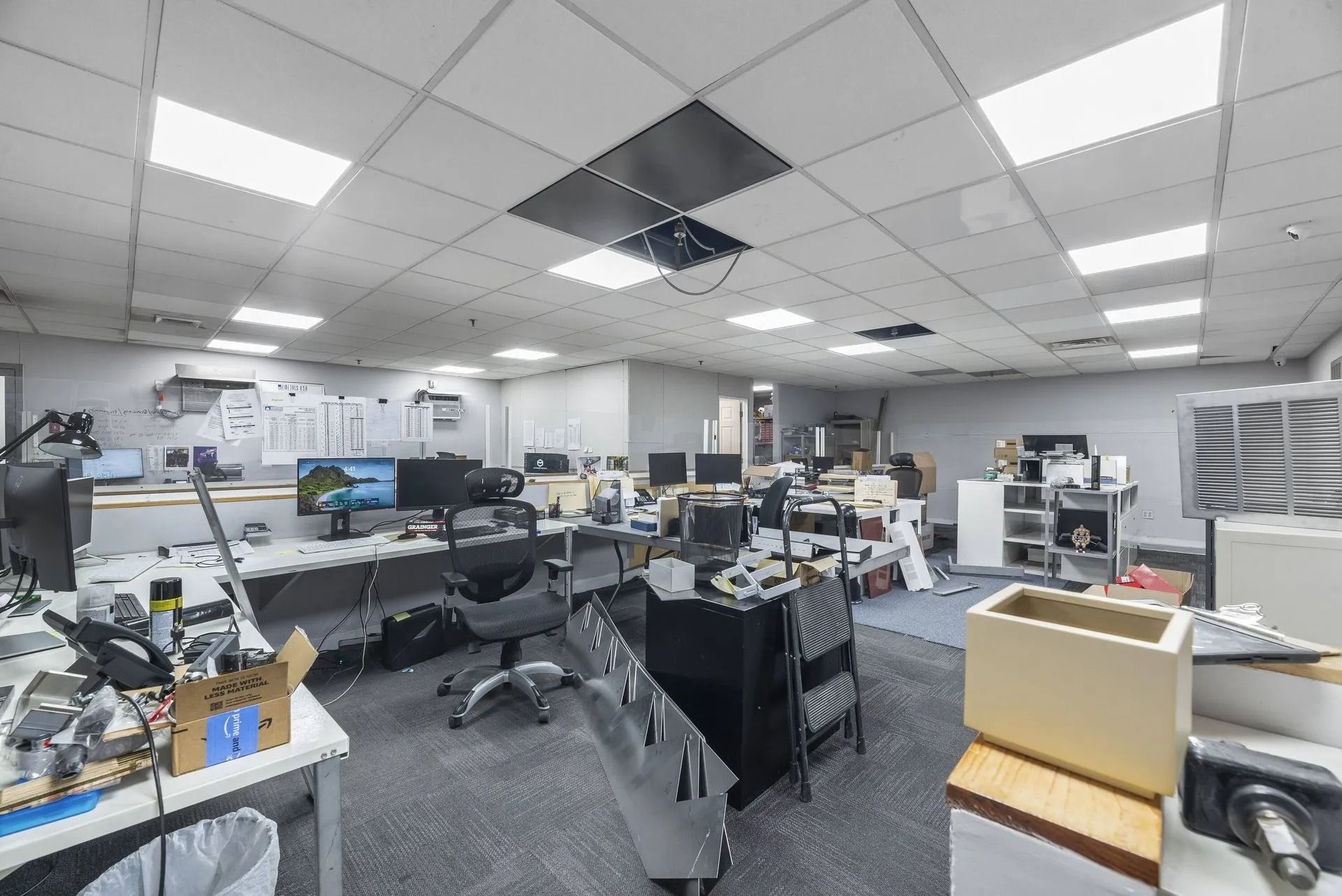 Open-plan office with desks, computer monitors, chairs, and fluorescent ceiling lights