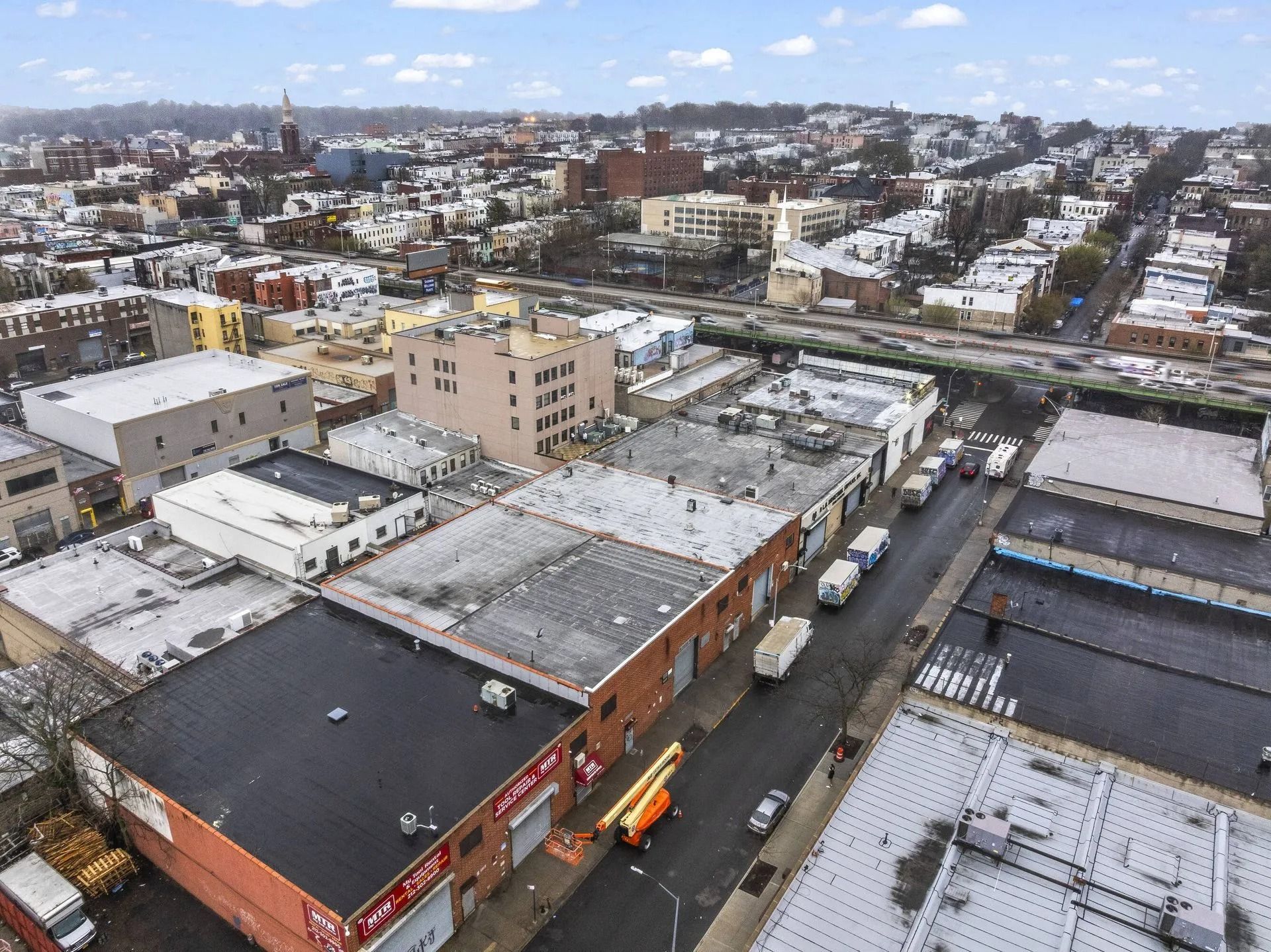 Aerial view of a snowy urban neighborhood with low-rise buildings, streets, and parked cars under a cloudy sky