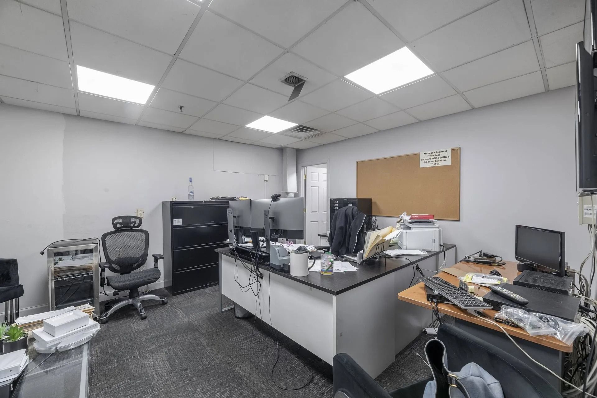 Office workspace with desks, computers, chairs, and a corkboard in a white-walled room.