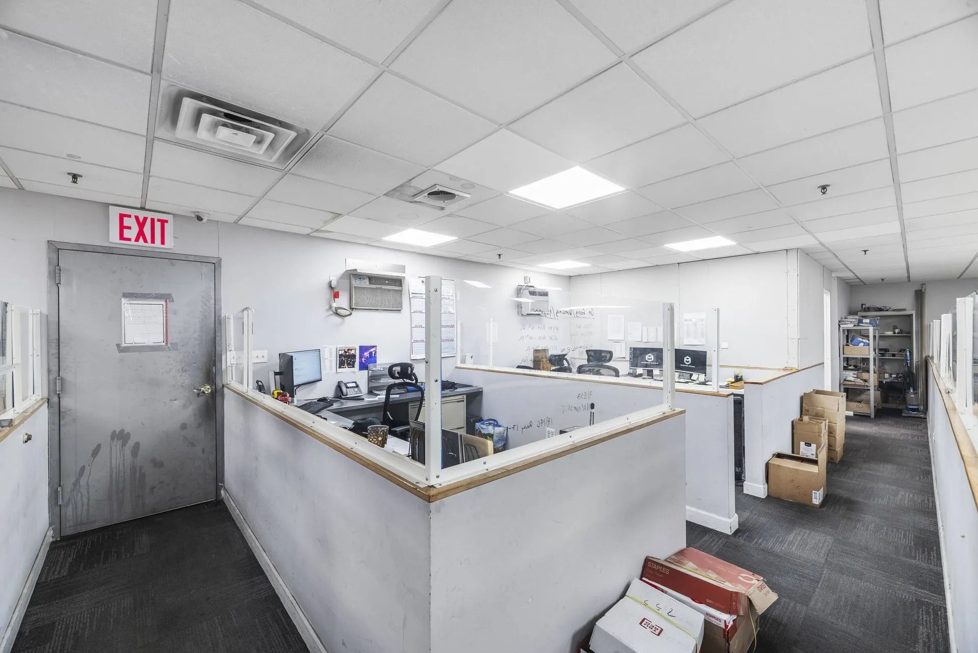 Open office with cubicles, computers, and an exit door under fluorescent lights.