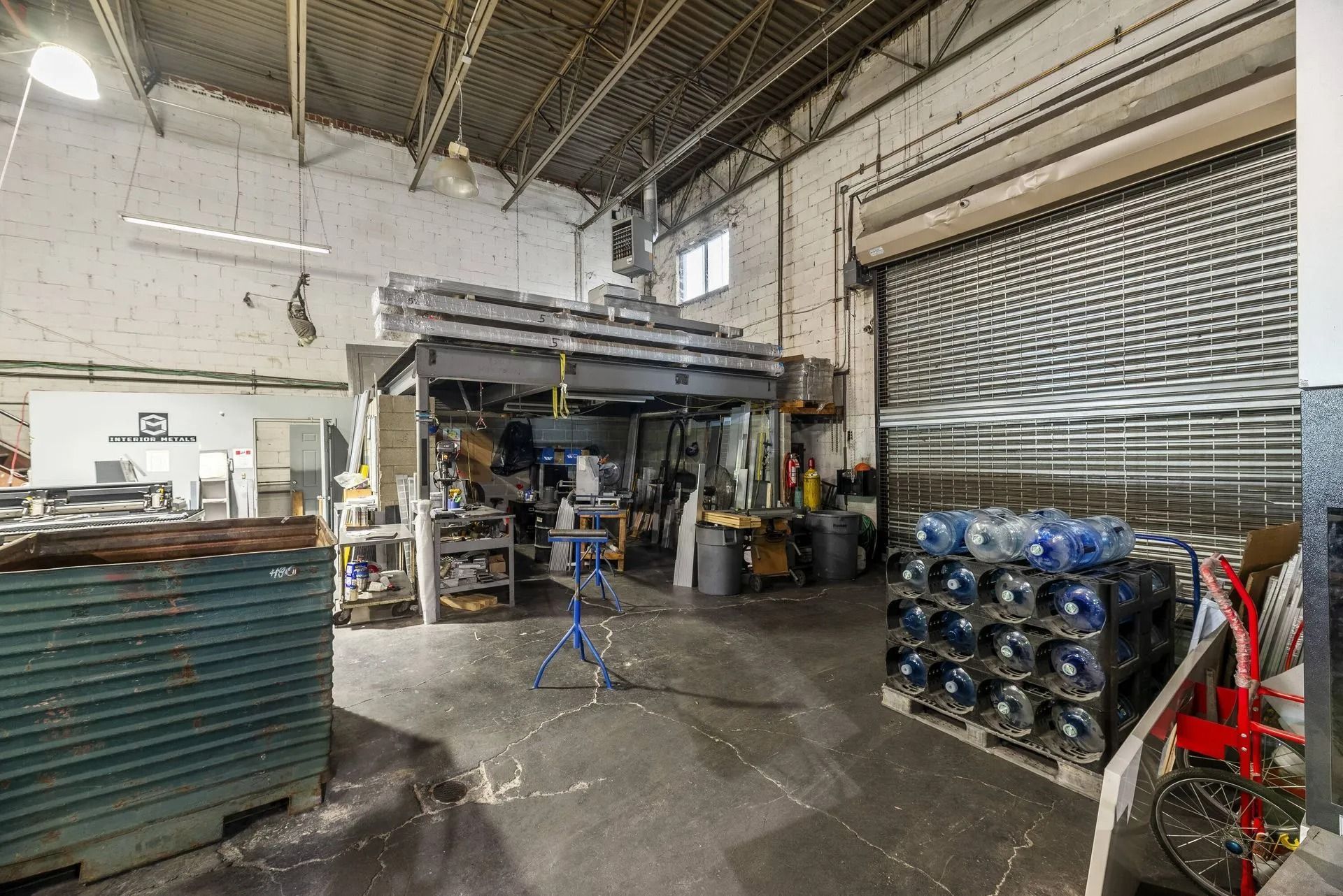 Industrial warehouse interior with machinery, stacked metal parts, and a partially open roll-up door