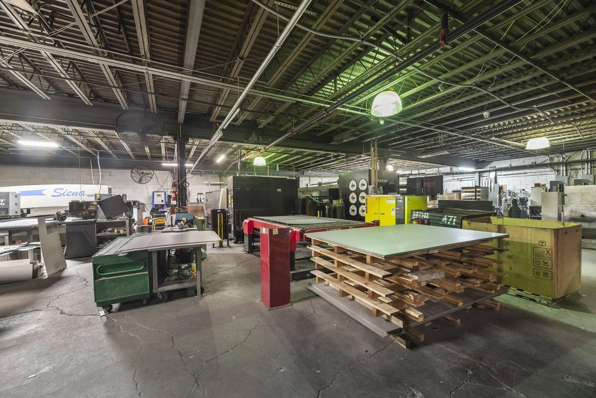 Industrial workshop interior with stacked pallets, machinery, and high metal ceiling lights
