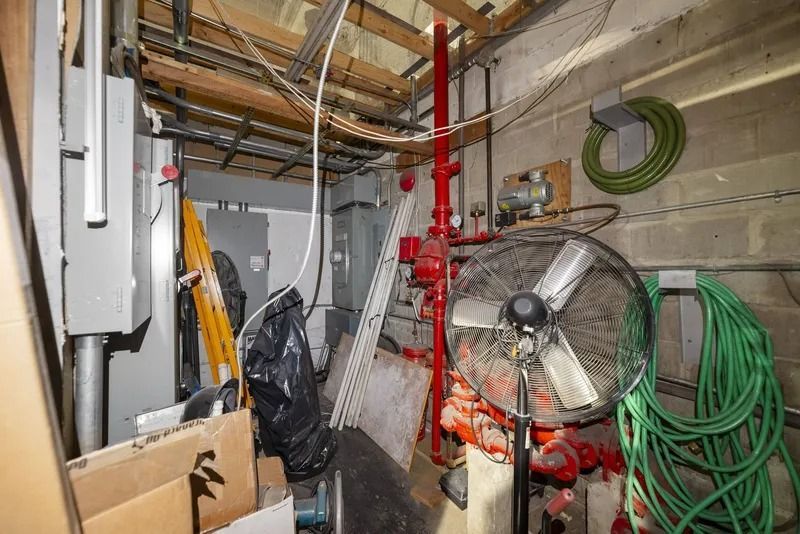 Cluttered utility room with pipes, hoses, fan, and stored tools against concrete walls.