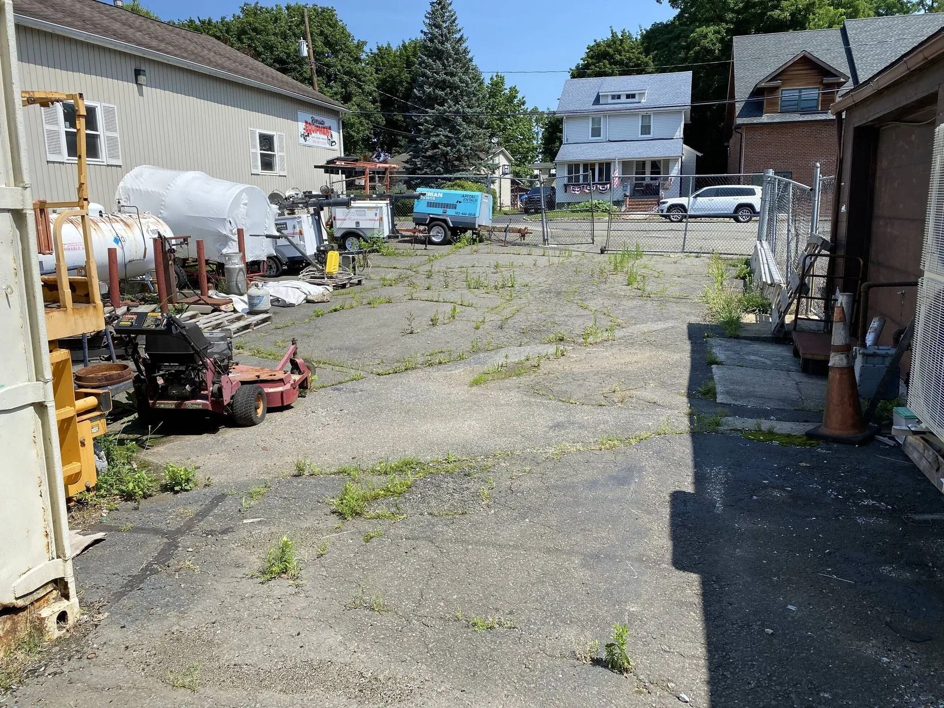 Overgrown paved lot between buildings with tools, tanks, and parked vehicles under a clear blue sky