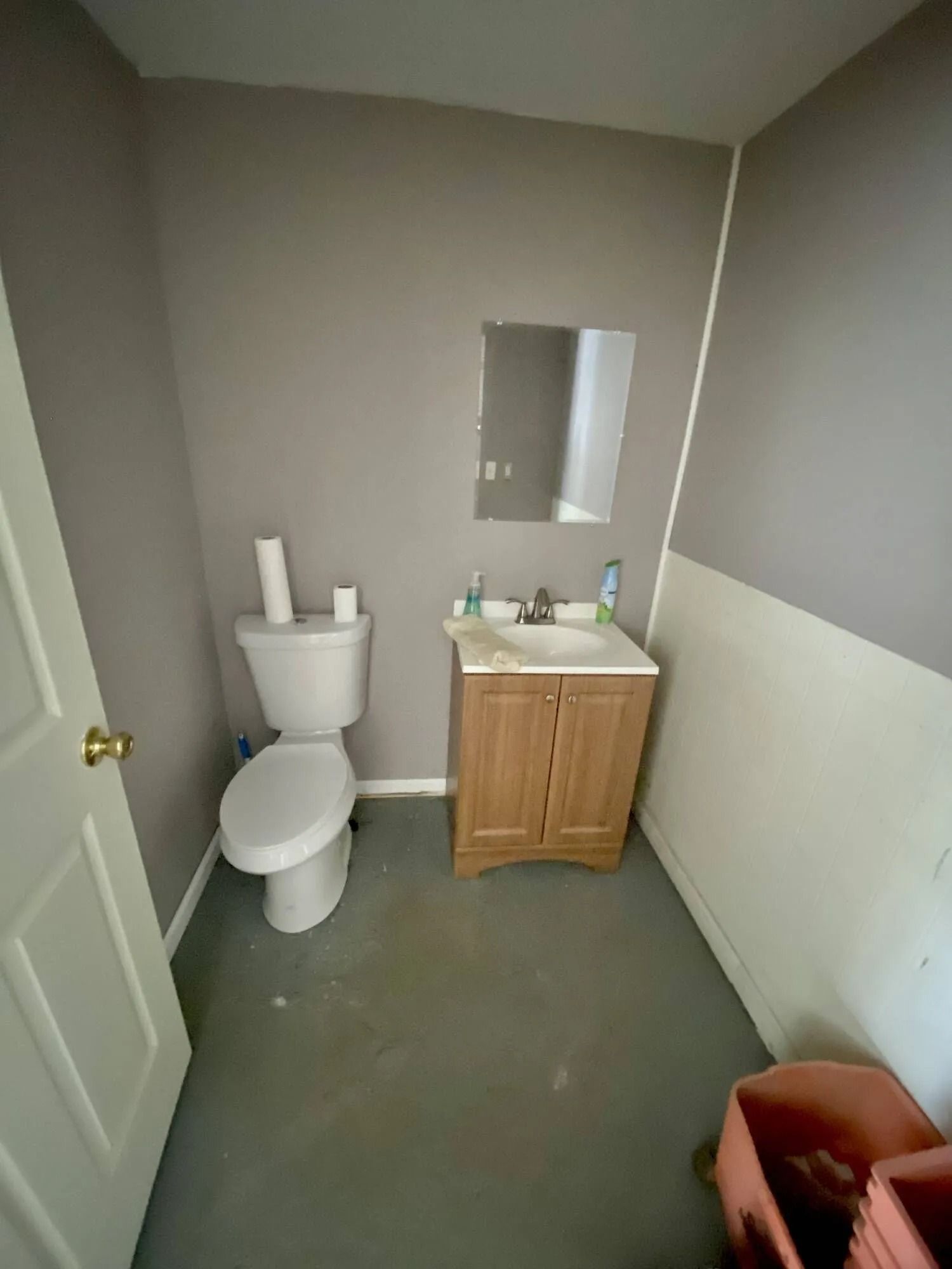Small bathroom with toilet, sink, mirror, gray walls, and a trash bin in the corner.
