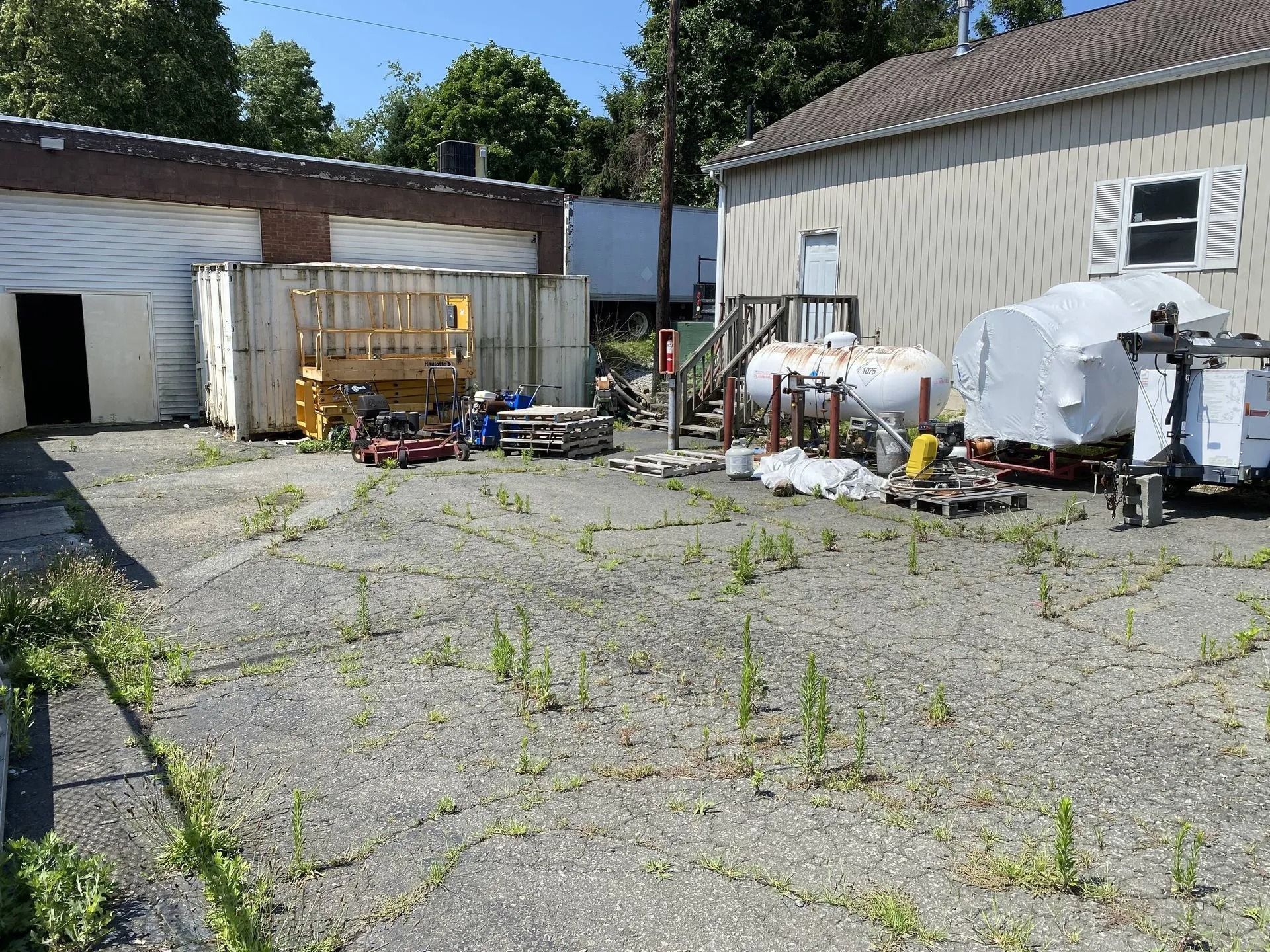 Outdoor industrial yard with scattered equipment, white tanks, and a garage beside a beige building.