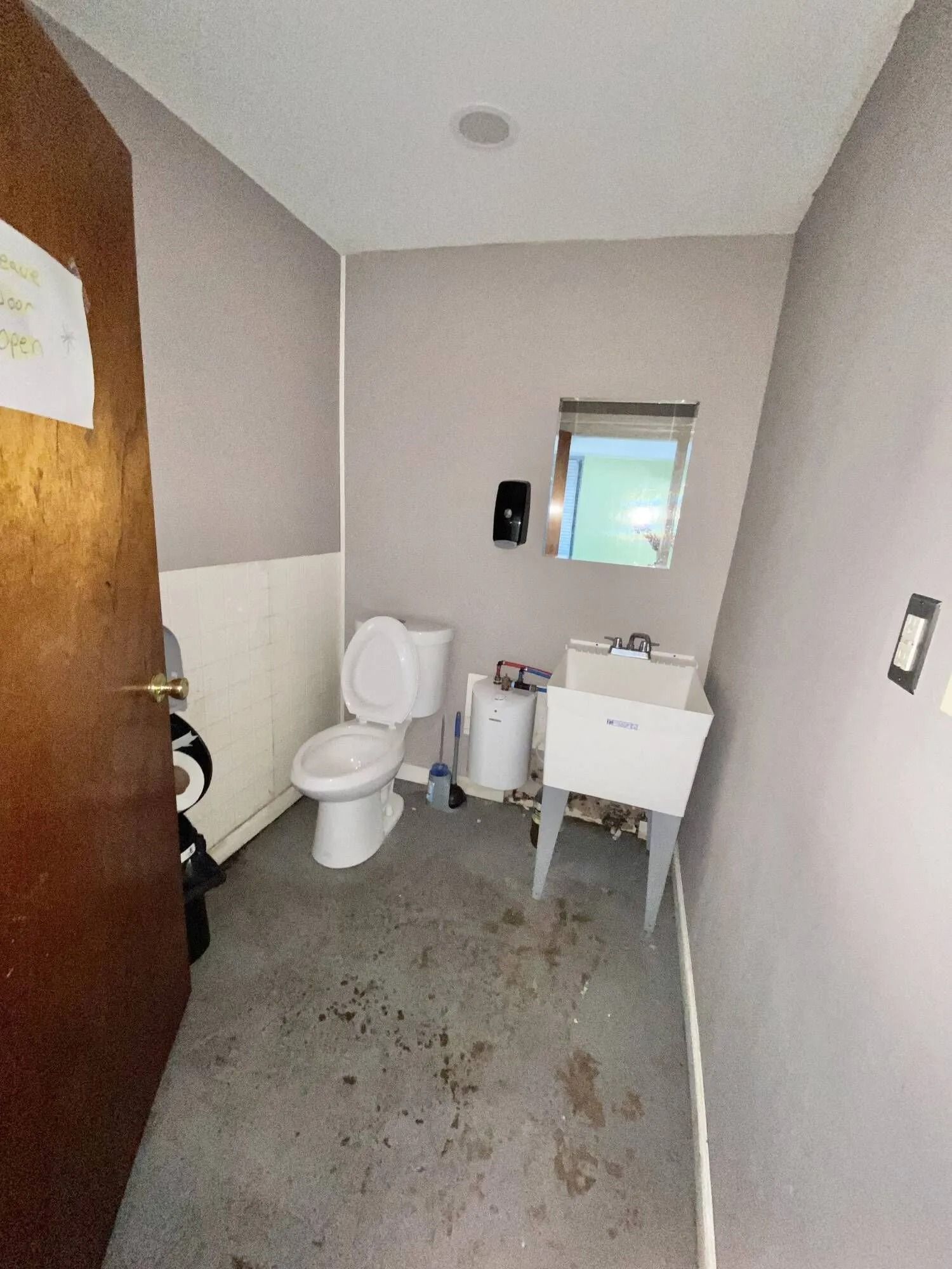 Small restroom with toilet, sink, and mirror; dirty floor and open wooden door.