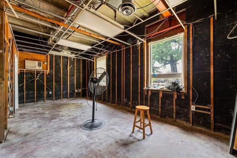 Unfinished room with exposed studs, ceiling fan, window, pedestal fan, and a stool on a concrete floor.