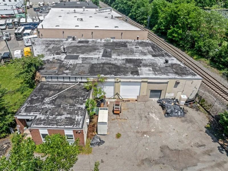 Aerial view of an industrial warehouse yard with flat roofs, paved space, trees, and railroad tracks nearby