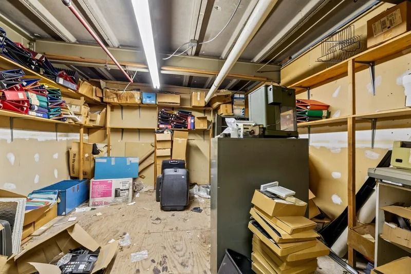 Cluttered storage room with shelves of boxes, papers, and a black shredder under fluorescent lights.