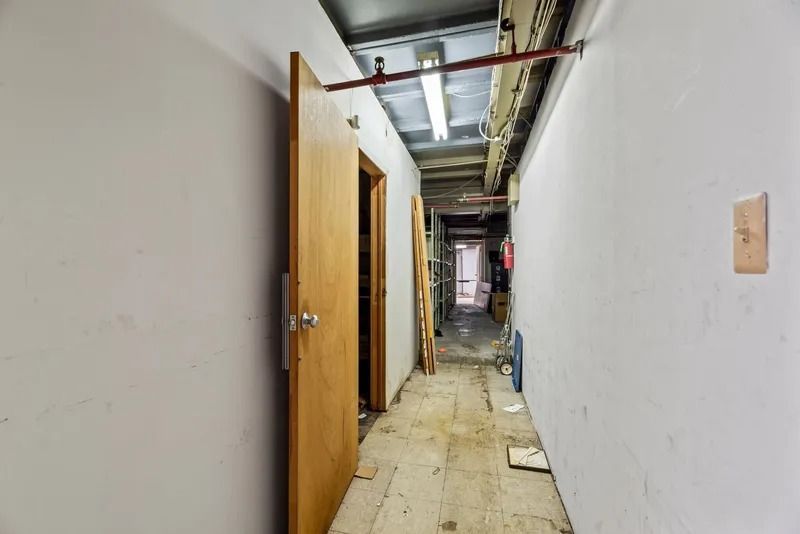 Narrow unfinished hallway with exposed ceiling pipes, open wooden door, and construction debris on the floor