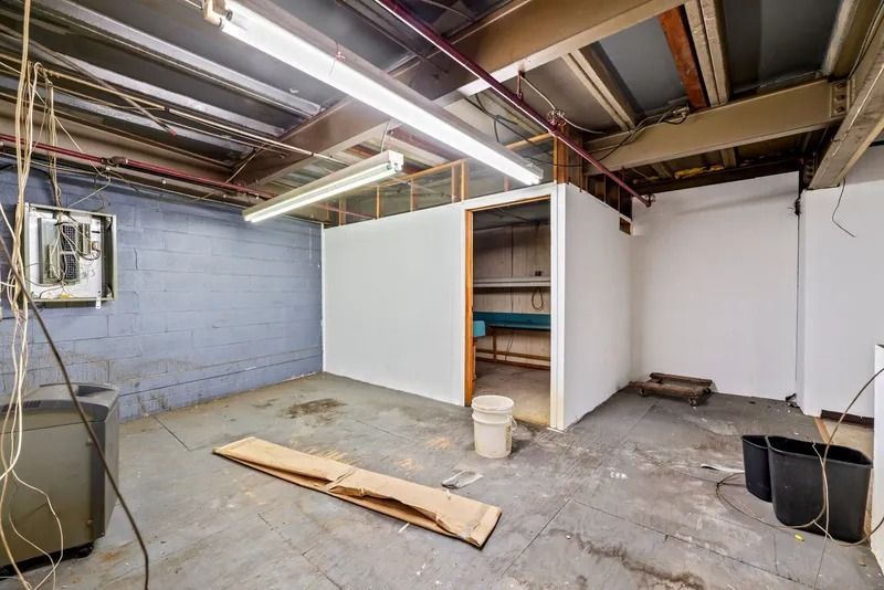 Empty basement with exposed ceiling, concrete floor, white walls, shelves, and buckets during renovation.
