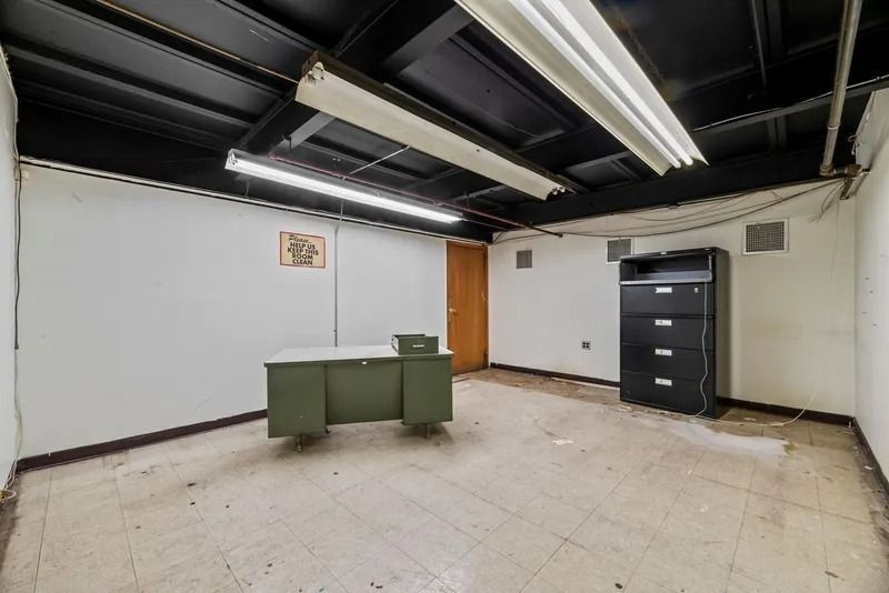 Empty basement room with fluorescent lights, white walls, a green cabinet, and a black filing cabinet.