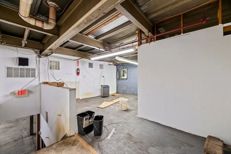 Basement interior with exposed pipes, white walls, concrete floor, and a trash bin near stairs
