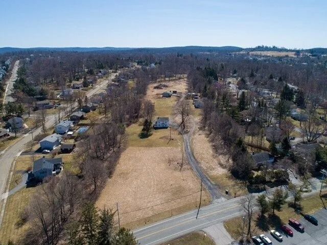 Aerial view of a suburban neighborhood with roads, houses, trees, and open grassy lots.