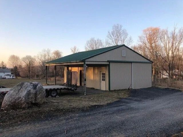 Small metal shed with green roof and covered porch beside a gravel drive at sunset