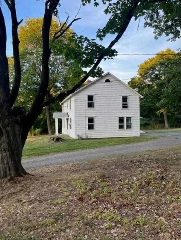 White two-story house by a gravel road, framed by tall trees in autumn.
