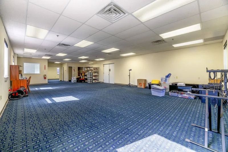Empty carpeted office room with drop ceiling, fluorescent lights, and scattered equipment along the walls