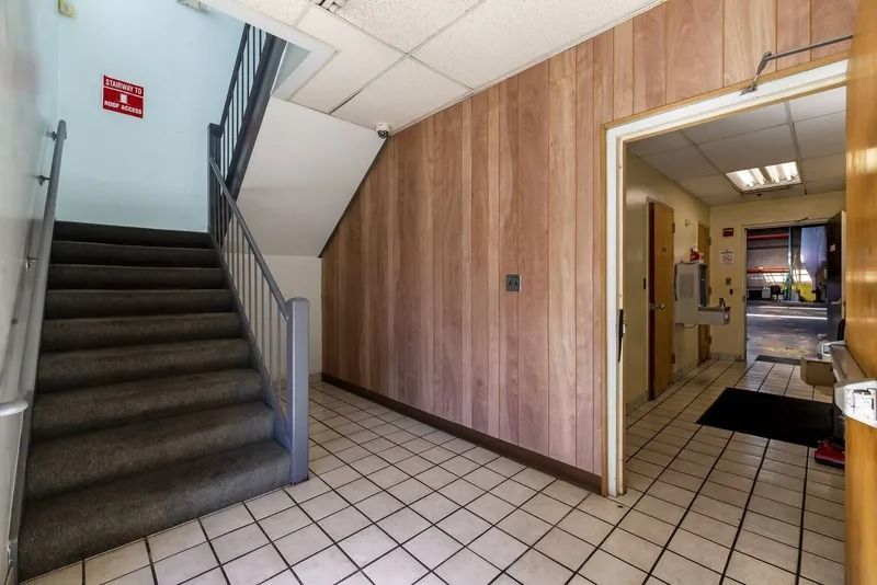 Indoor stairwell with gray carpeted stairs, wood-paneled wall, and a tiled hallway opening to an office area.