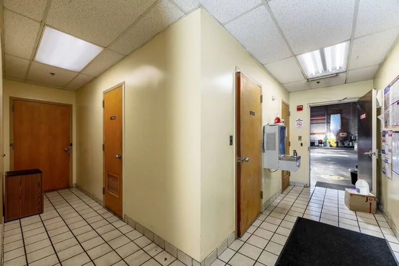 Empty office hallway with beige walls, tiled floor, wooden doors, and fluorescent ceiling lights