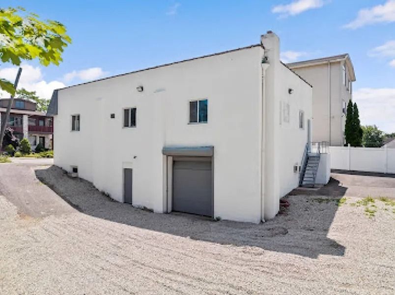White stucco building with small windows, a garage door, and an exterior staircase on a gravel lot