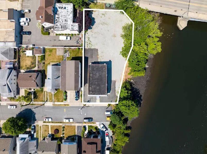 Aerial view of a waterfront property with a house, driveway, parked cars, and trees beside dark water.