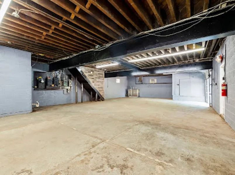 Empty unfinished basement with exposed beams, concrete floor, staircase, and utility doors.