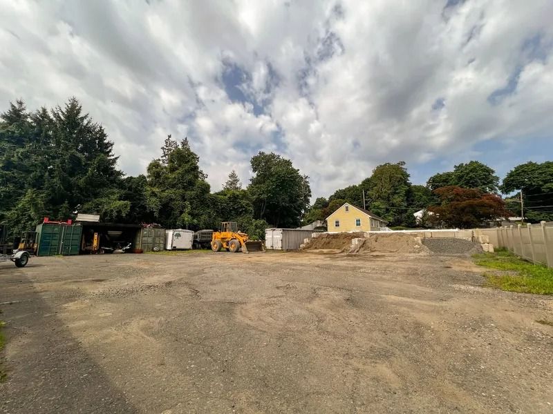 Wide empty gravel lot with a small yellow house, trees, and cloudy sky in the background