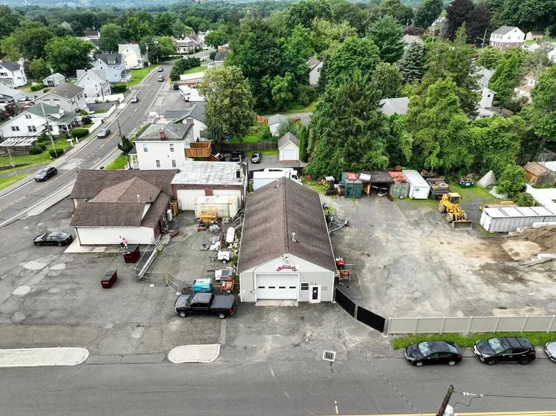 Aerial view of a small commercial lot with a long building, parked vehicles, and surrounding trees and houses.