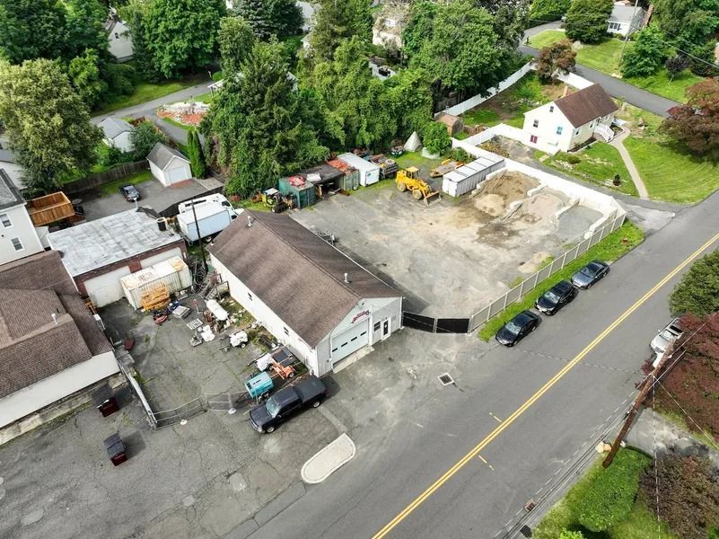 Aerial view of a rural yard with buildings, parked cars, and trees beside a road.
