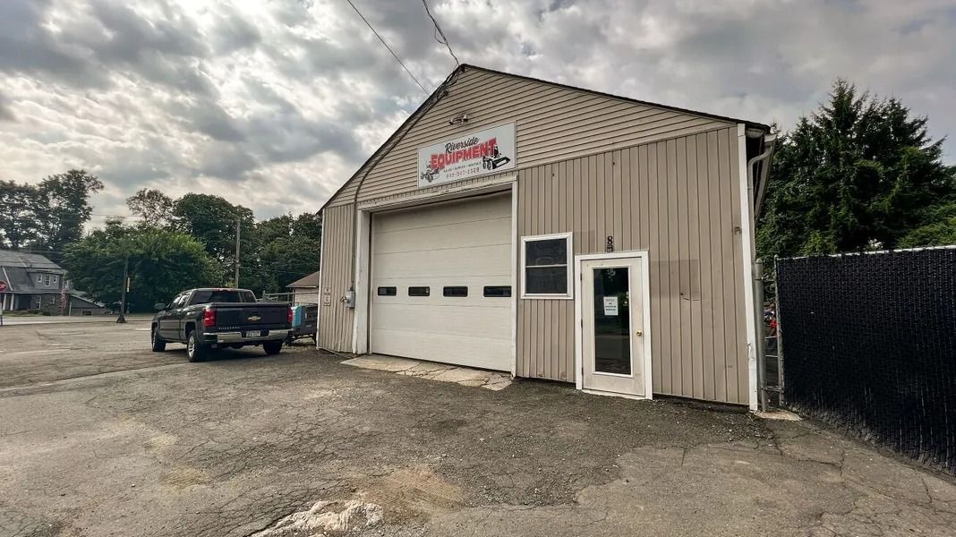 Tan metal garage with white bay doors, sign above entrance, and a black pickup truck parked outside under cloudy sky