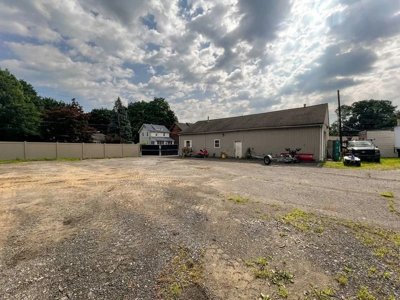 Gravel lot beside a beige building under a cloudy sky, with a truck, trailer, and parked vehicles.