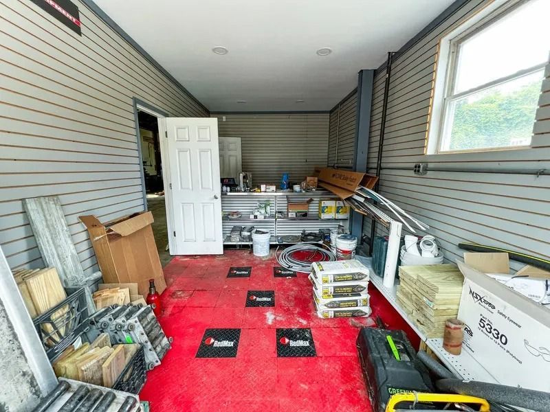 Cluttered covered porch or garage with red carpet, stacked boxes, shelves, and a white door at the back